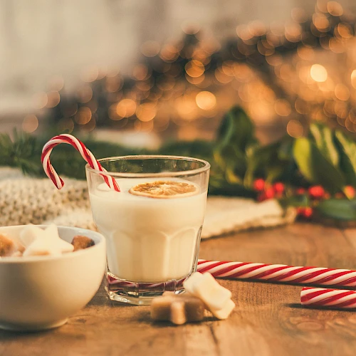 A cozy holiday scene with a glass of warm milk, candy cane, and sugar cookies on a wooden table, twinkling lights in the background.