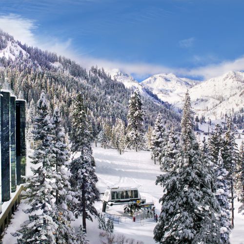 A snowy mountain landscape with a modern glass building reflecting the scenery, surrounded by snow-covered trees under a blue sky.