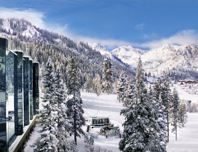 A snowy mountain landscape with a modern glass building reflecting the scenery, surrounded by snow-covered trees under a blue sky.