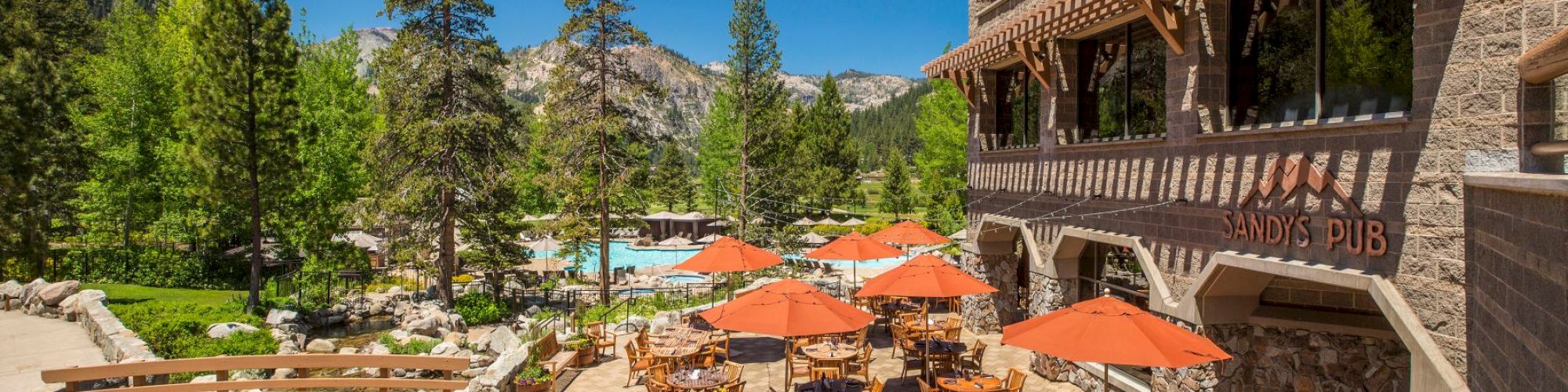 An outdoor dining area with tables, chairs, and orange umbrellas is set amid a lush, mountainous landscape near a building marked 