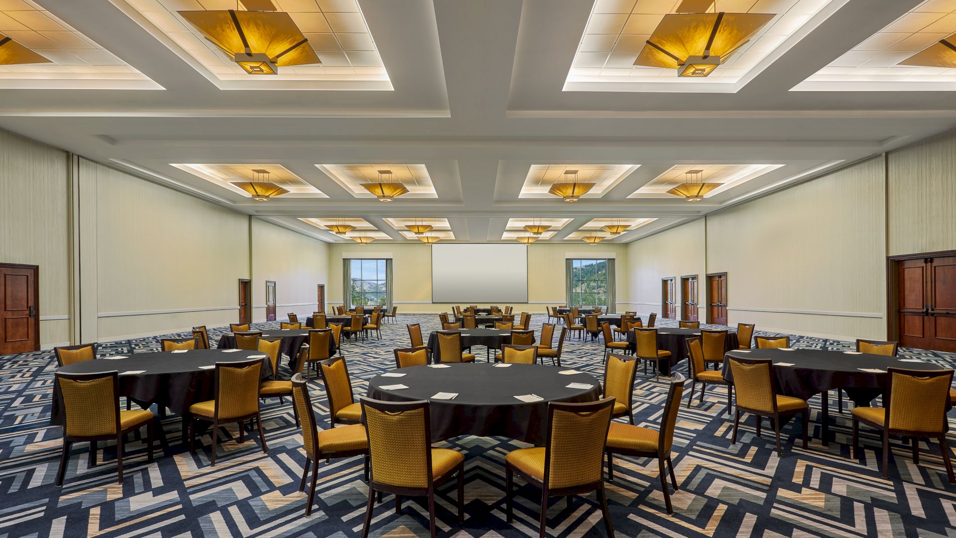 A spacious, well-lit conference room with round tables and yellow cushioned chairs, patterned carpet, and modern ceiling lights.