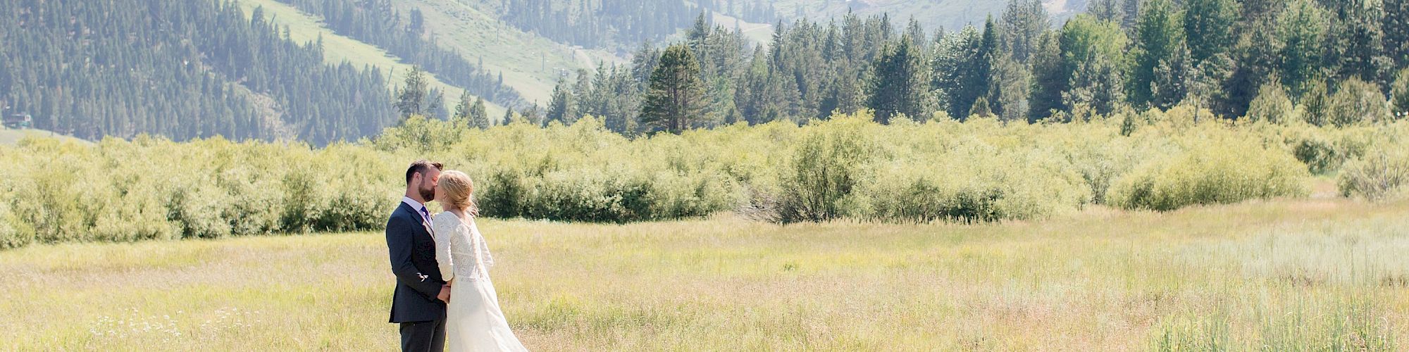 A couple in wedding attire stands together in a grassy field with a scenic mountain backdrop. They appear to be sharing a romantic moment.