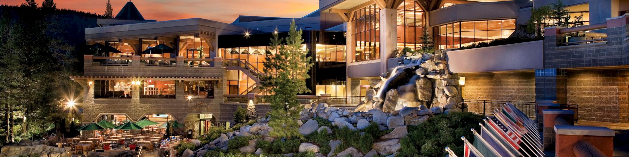 A modern building with large windows and outdoor seating in a mountainous area at sunset, surrounded by flags from various countries.