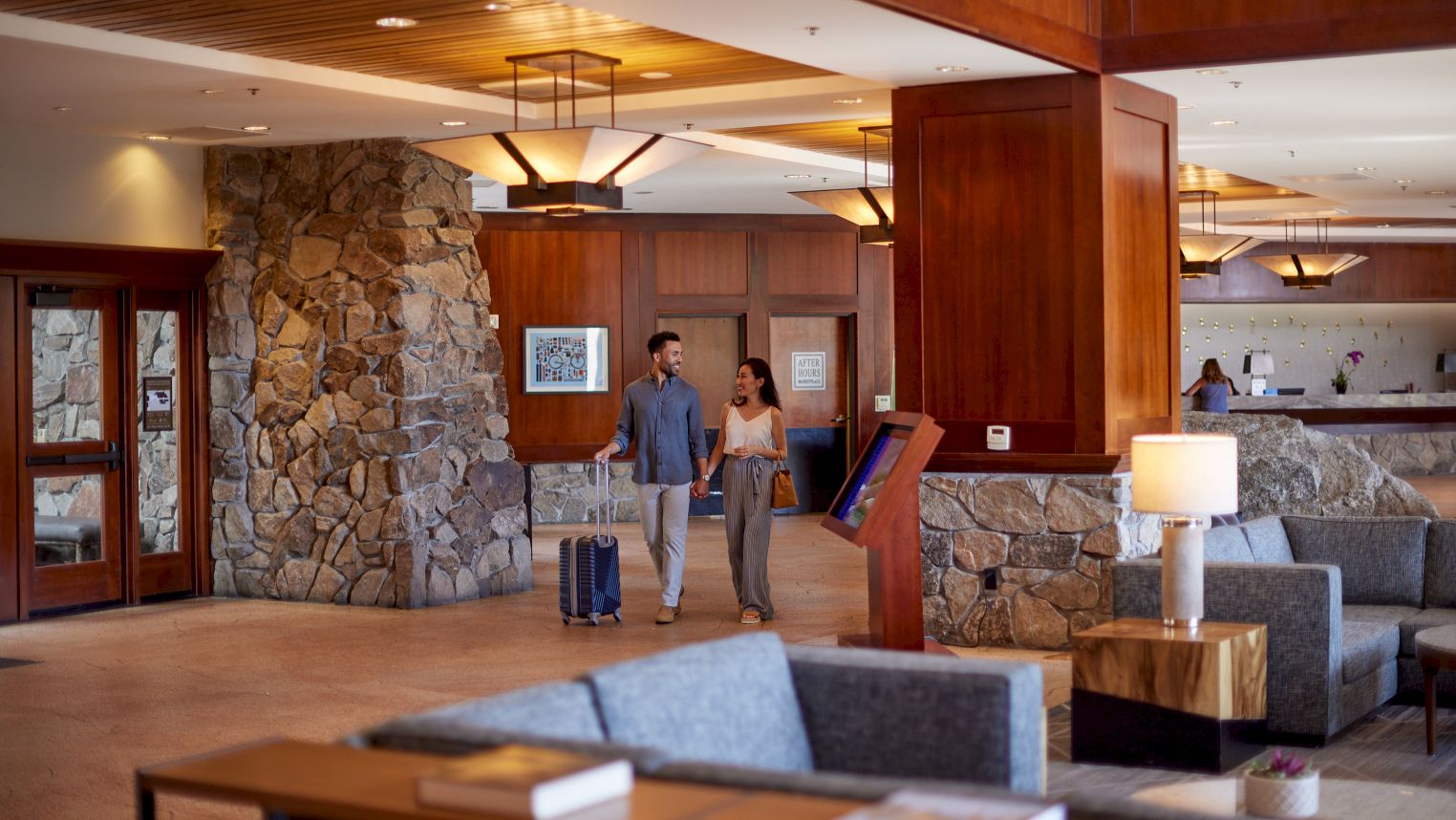 Two people walk through a hotel lobby with suitcases, past a reception desk with staff and seating area.