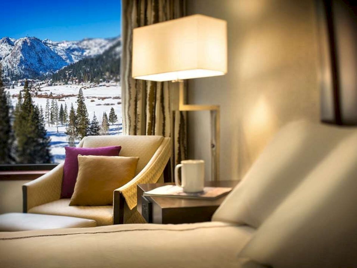 A cozy hotel room with a mountain view, featuring a bed, a chair with cushions, a side table, and a lamp. Snowy landscape is seen outside.