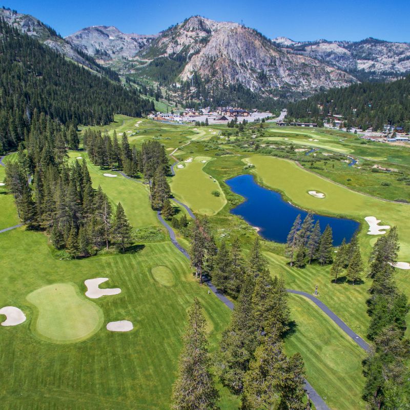 The image shows a scenic golf course surrounded by trees and mountains, with a pond, fairways, and sand traps. A building is visible in the foreground.