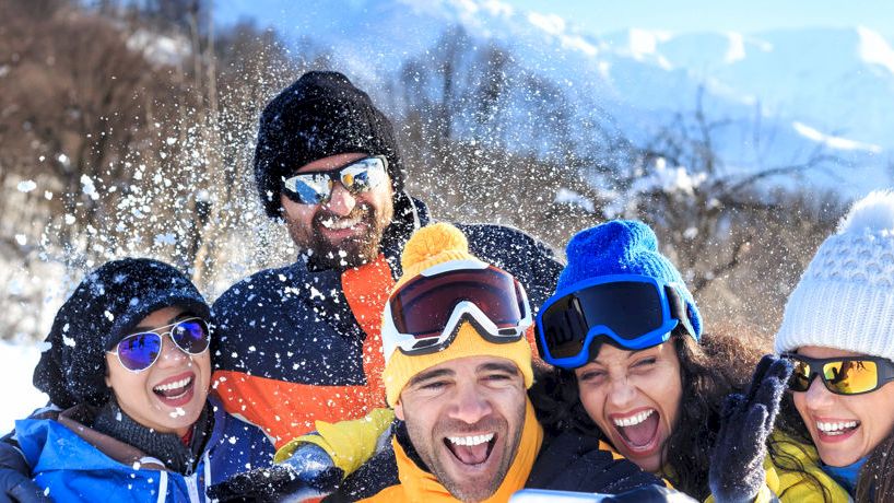 A group of five people in winter clothing are laughing and enjoying the snowy outdoors, with mountains and trees in the background.