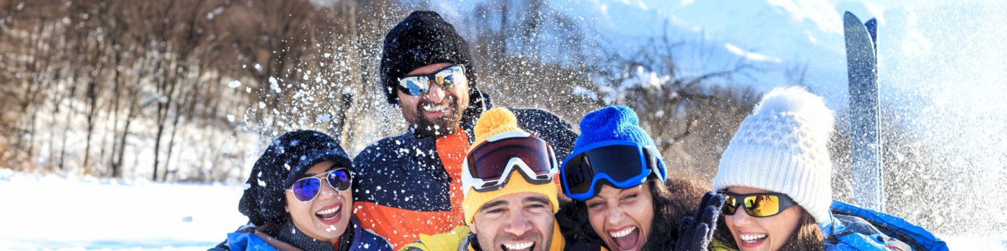 A group of five people in winter clothing are laughing and enjoying the snowy outdoors, with mountains and trees in the background.