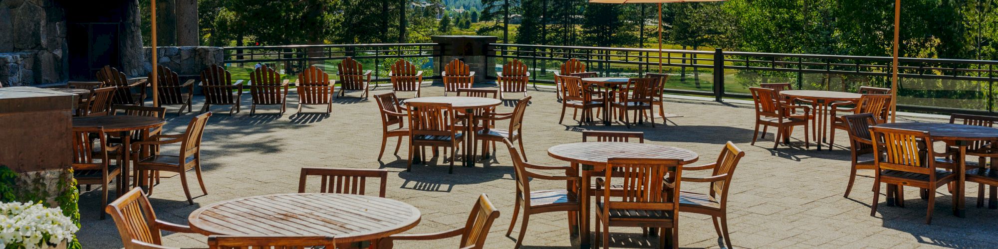 An outdoor patio with wooden tables and chairs, some with umbrellas, surrounded by trees and mountains in the background on a sunny day.