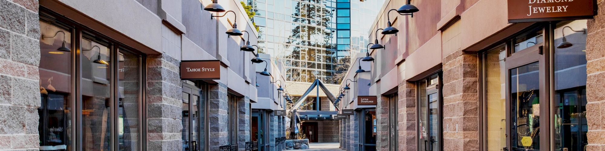 An outdoor shopping walkway with glass storefronts on both sides, leading to a modern glass building in the background.