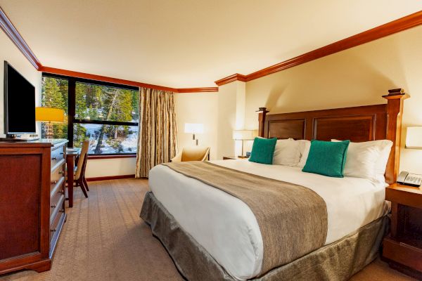 A cozy hotel room with a king-size bed, green pillows, wooden furniture, a TV, a desk with a chair, and a window showing a scenic outdoor view.