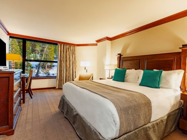 A cozy hotel room with a king-size bed, green pillows, wooden furniture, a TV, a desk with a chair, and a window showing a scenic outdoor view.
