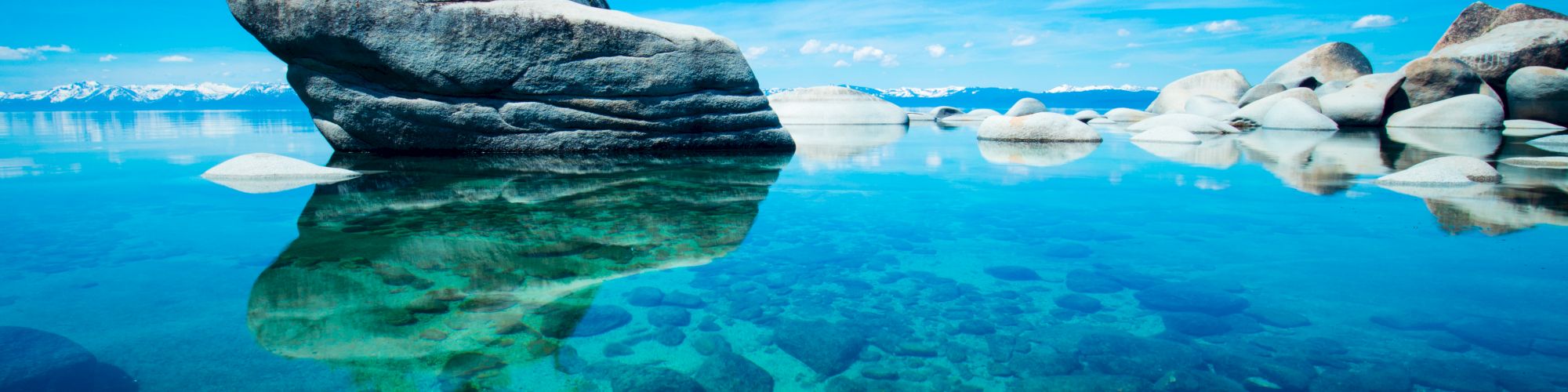 A serene lake with clear, turquoise water, showcasing a large boulder and smaller rocks amid a scenic background of mountains and a bright blue sky.