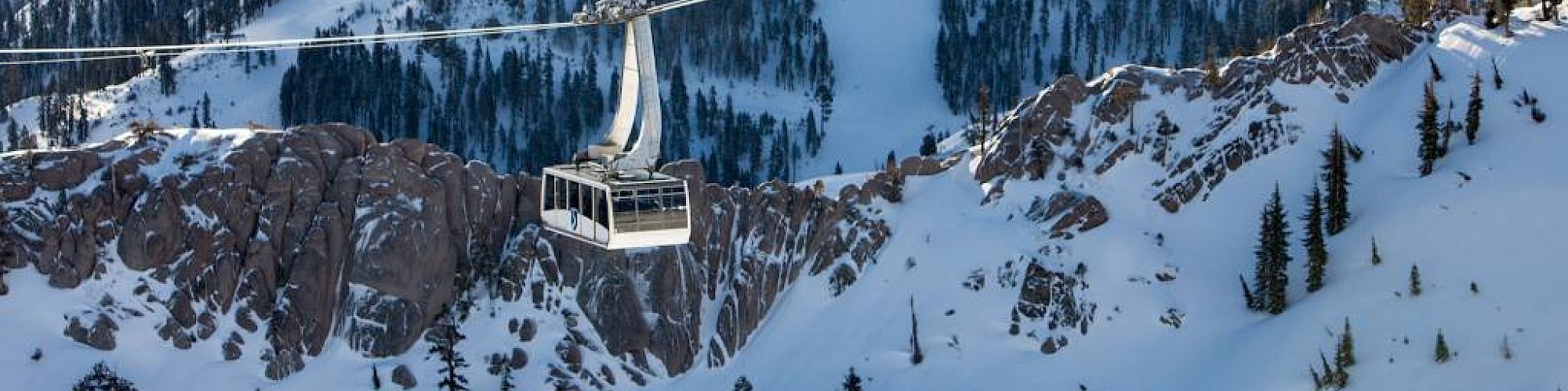 A scenic snowy mountain landscape featuring a cable car suspended between peaks, surrounded by forested areas and with a lake in the background.