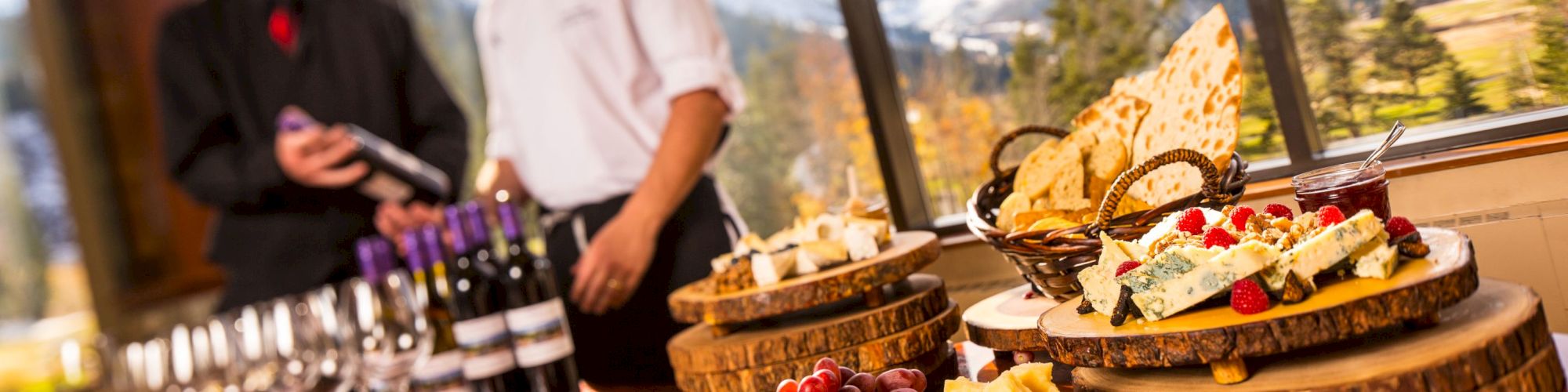 A table with assorted cheeses, fruits, and bread is set up in front of a window with a scenic mountain view. Two people are conversing behind the table.