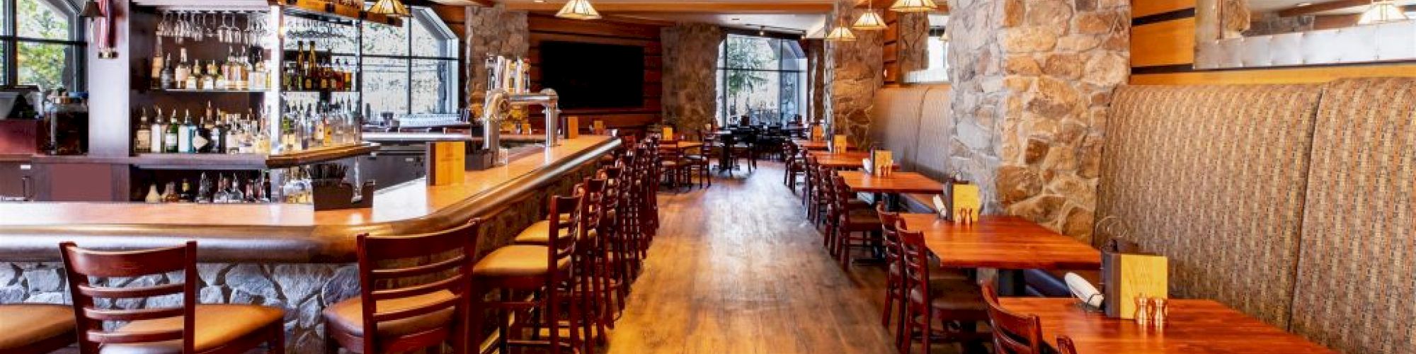 The image shows an empty restaurant with wooden tables and chairs, a well-stocked bar on the left, and a rustic stone wall on the right, under warm lighting.