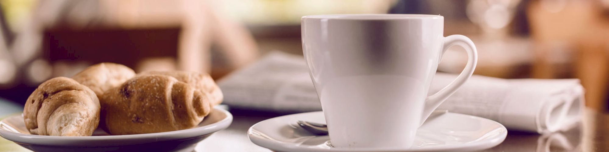 A white coffee cup on a saucer sits on a table next to a plate of croissants and a rolled-up napkin, with a blurry background.