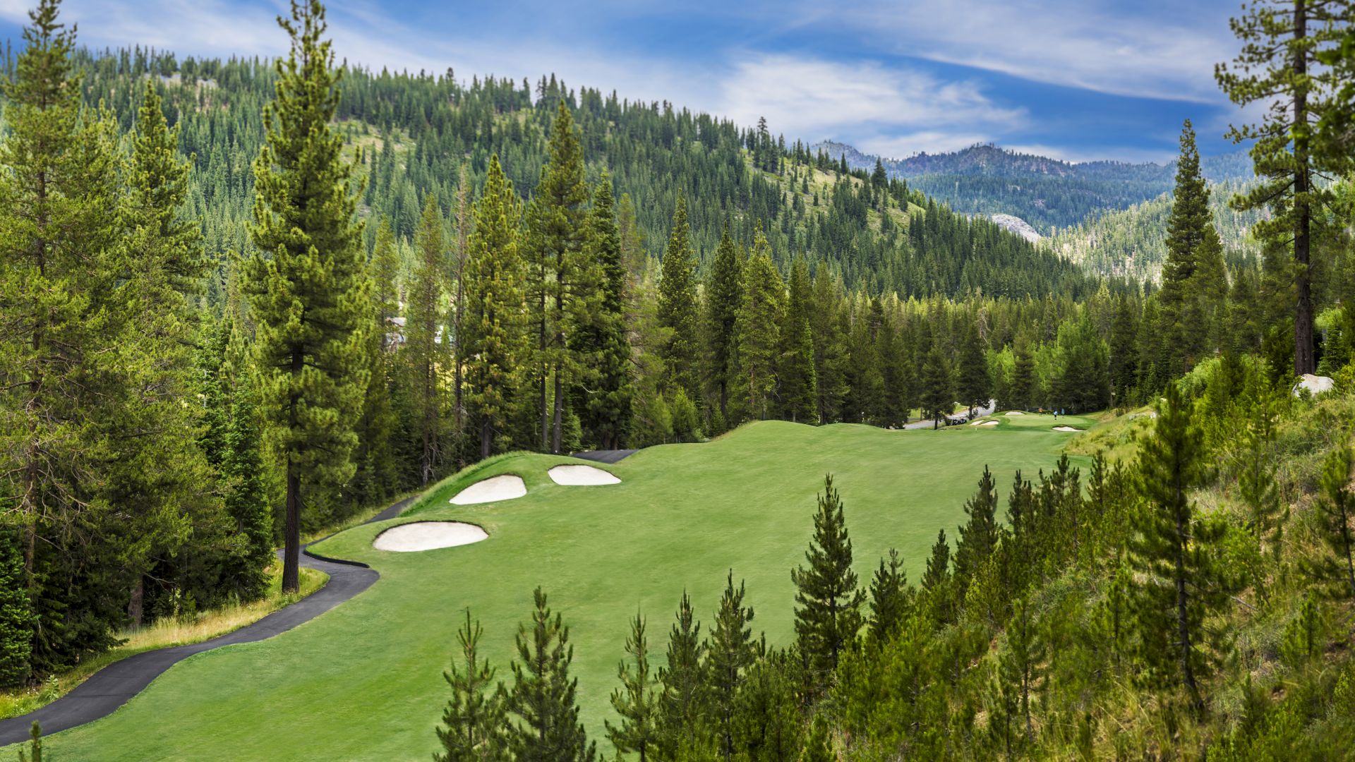 A scenic golf course with lush greens, sand bunkers, and a winding path, surrounded by tall pine trees and forested mountains under a blue sky.