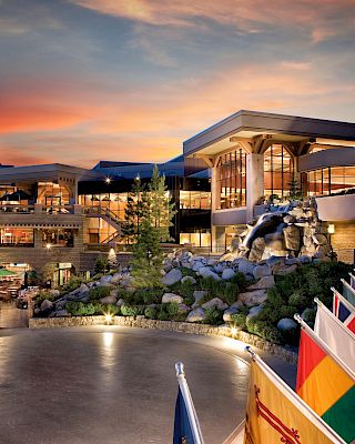 A modern building at sunset with international flags in the foreground, surrounded by trees and rocky landscaping.