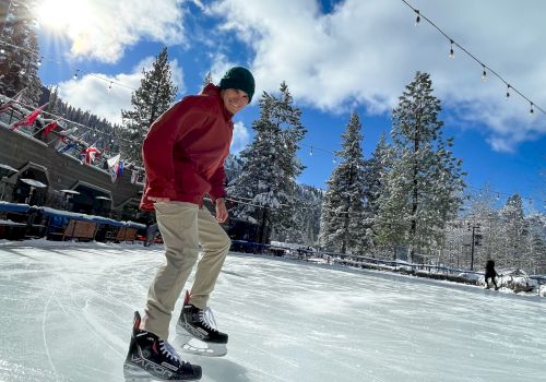 A person wearing a red jacket and beige pants glides on ice at a snowy rink, with string lights, trees, and a blue sky in the background.