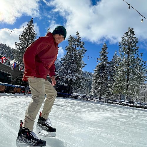 A person wearing a red jacket and beige pants glides on ice at a snowy rink, with string lights, trees, and a blue sky in the background.