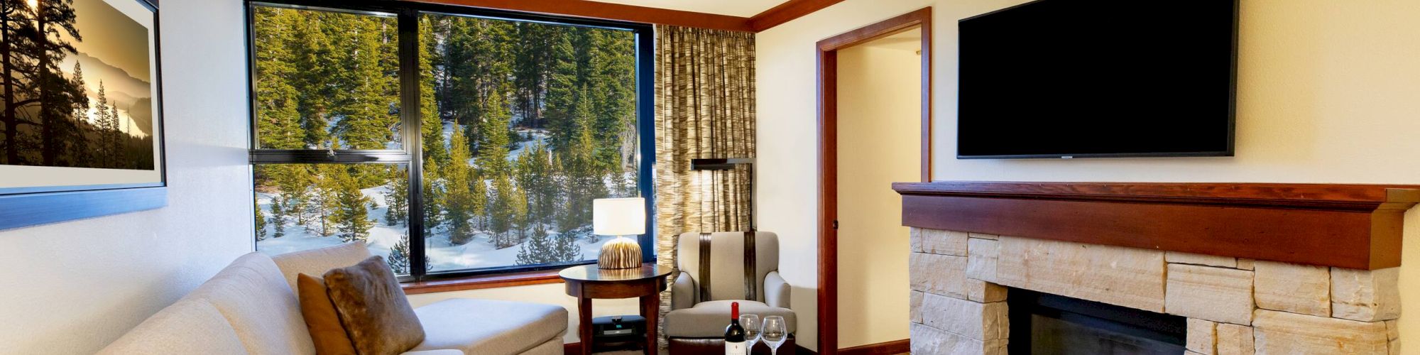 A cozy living room with a beige sofa, fireplace, wall-mounted TV, round coffee table, and large window overlooking trees.