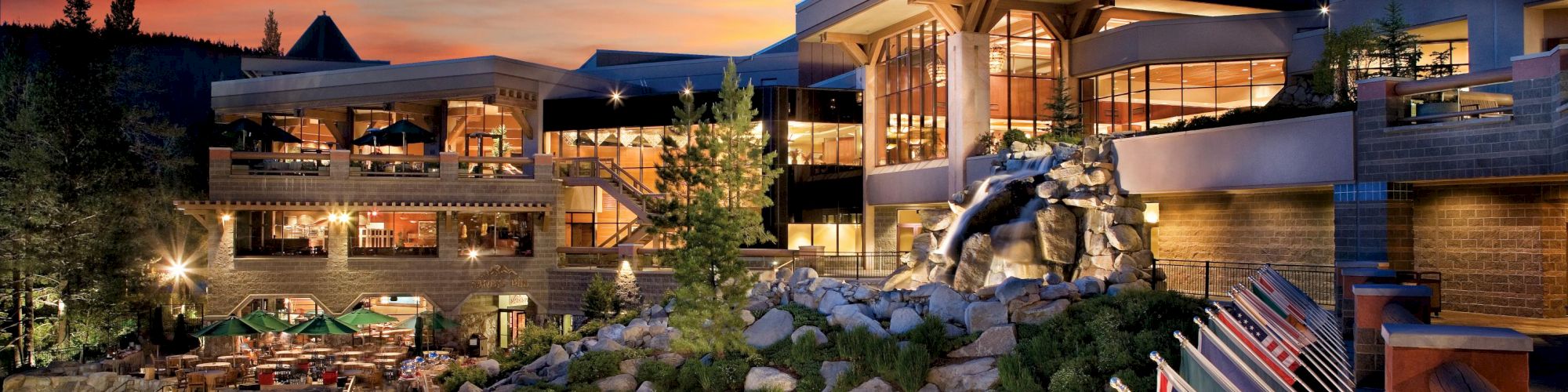 The image shows a beautiful building at sunset, surrounded by flags from different countries, with trees, a statue, and rocky landscaping.