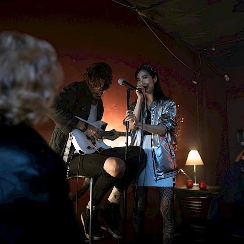 Two musicians perform in a cozy dim bar; a guitarist sits while a woman sings into a mic, warmly lit by a lamp, with a small audience watching.