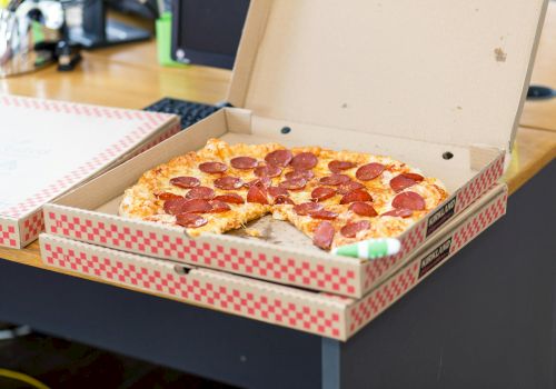A pepperoni pizza in an open box on a desk, with another box nearby; casual office or workstation setting.