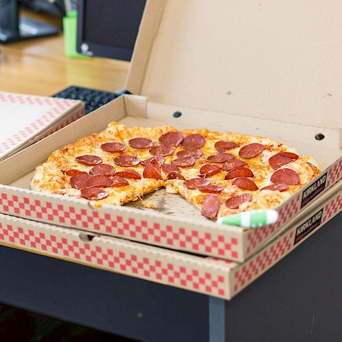 A pepperoni pizza in an open box on a desk, with another box nearby; casual office or workstation setting.