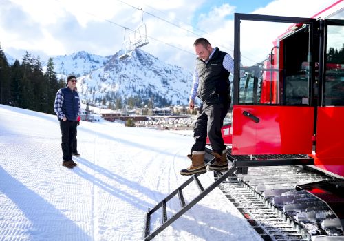 Two men in a snowy mountain area near a red snowcat; one climbs a ramp while the other watches.