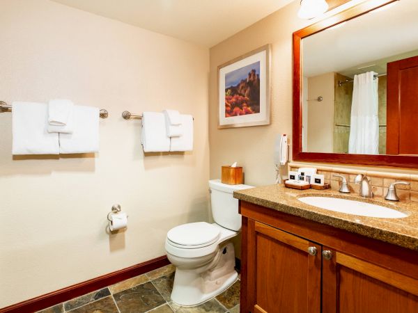 A clean bathroom with a toilet, towels, a sink with a countertop, mirror, and artwork on the wall, featuring warm wood accents.