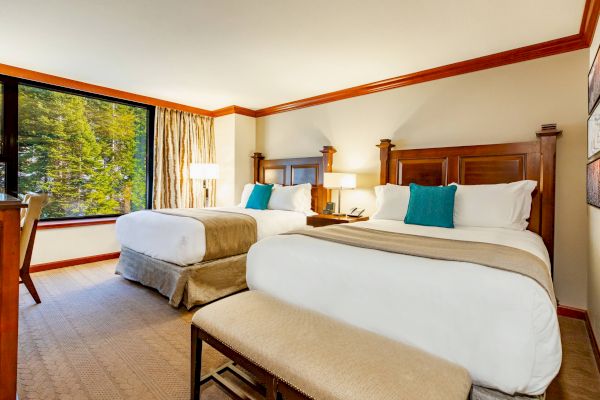 The image shows a hotel room with two double beds, wooden headboards, teal cushions, a large window, bedside lamps, and a beige carpet.