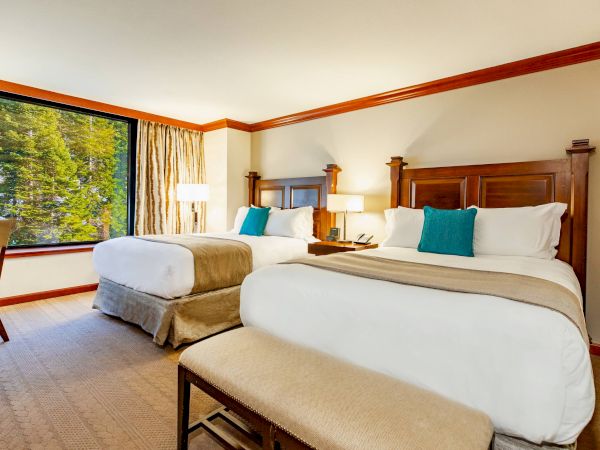 The image shows a hotel room with two double beds, wooden headboards, teal cushions, a large window, bedside lamps, and a beige carpet.