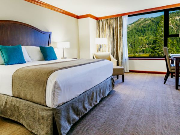 A hotel room with a large bed, pillows, and a scenic view from the window. There's a desk, chair, lamp, and a TV on a dresser.