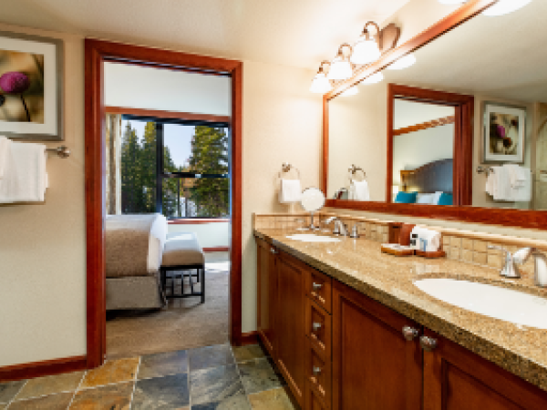 A bathroom with a double sink, large mirror, and art. Visible is a bed through the open doorway, framed by warm lighting.