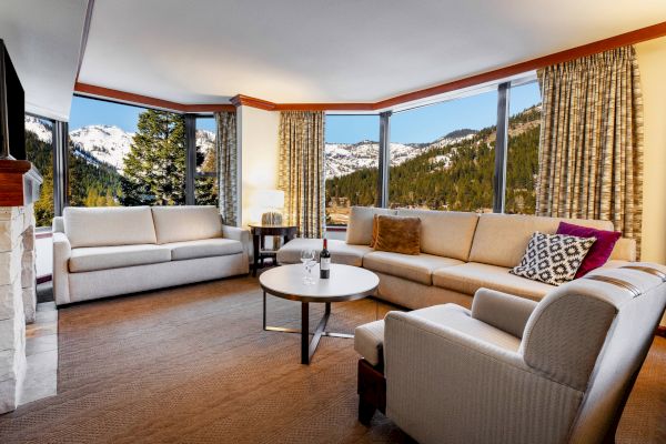 A cozy living room with beige furniture, large windows revealing a mountain view, a fireplace, and a round coffee table with a decor item.