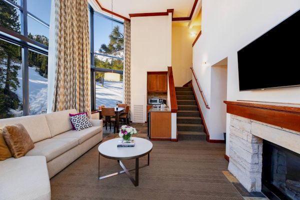 A modern living room with a beige sofa, coffee table, TV, fireplace, dining area, and stairs leading up. Large windows provide a scenic view outside.