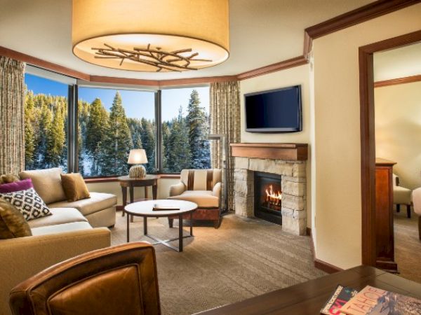 A cozy living room with a fireplace, sofa, chairs, and a TV, overlooking a snowy forest through large windows, adjoins a bedroom.
