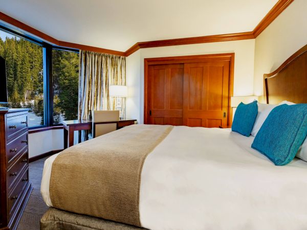 A cozy hotel room with a large bed, blue pillows, wooden furniture, and a scenic forest view through the window.