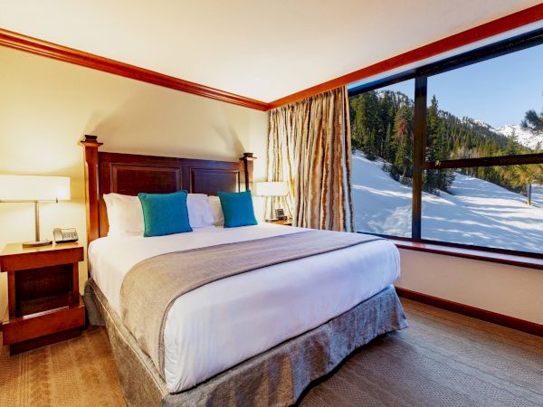 A cozy bedroom with a large bed, two bedside tables, lamps, and a window offering a snowy mountain view on a sunny day.
