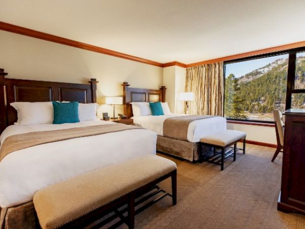 A hotel room with two beds, a large window with a scenic view, cushions, and a dresser with a lamp and TV.