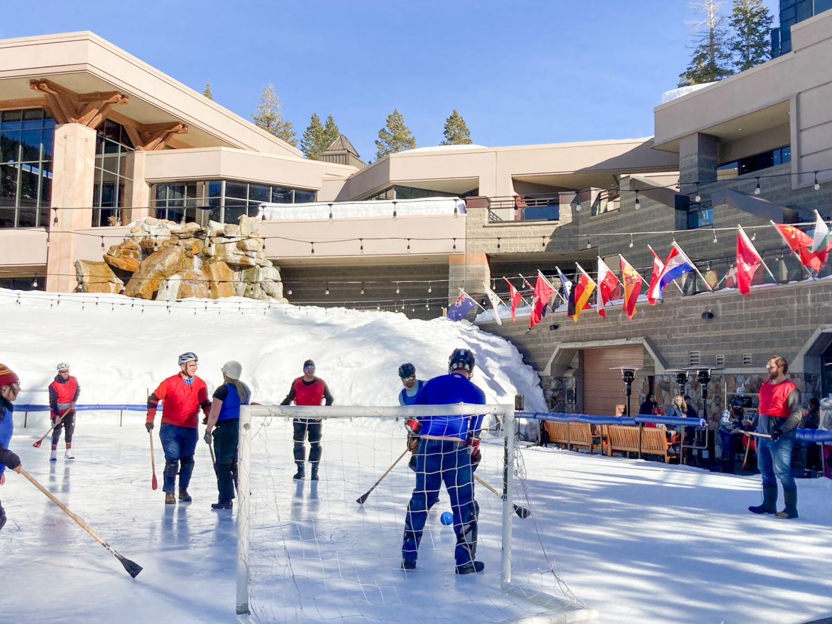 People play hockey on an outdoor rink at a sunny venue, with players in colorful gear and flags along the stands behind them.