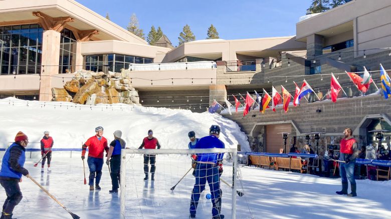 People play hockey on an outdoor rink at a sunny venue, with players in colorful gear and flags along the stands behind them.