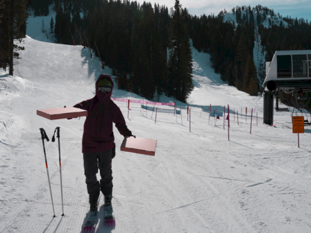 A person in ski gear holding a pink snowboard on a snowy slope, with trees, ski poles, and a chairlift in the background.