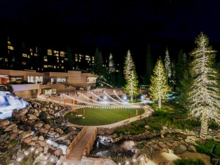 The image shows a beautifully lit outdoor area at night, featuring a manicured lawn, tall trees, string lights, and a building.