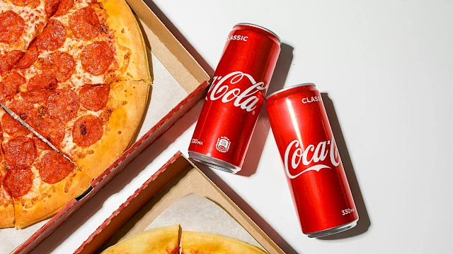 Two pizza slices in boxes with two red Coca-Cola cans beside them, set on a light background.