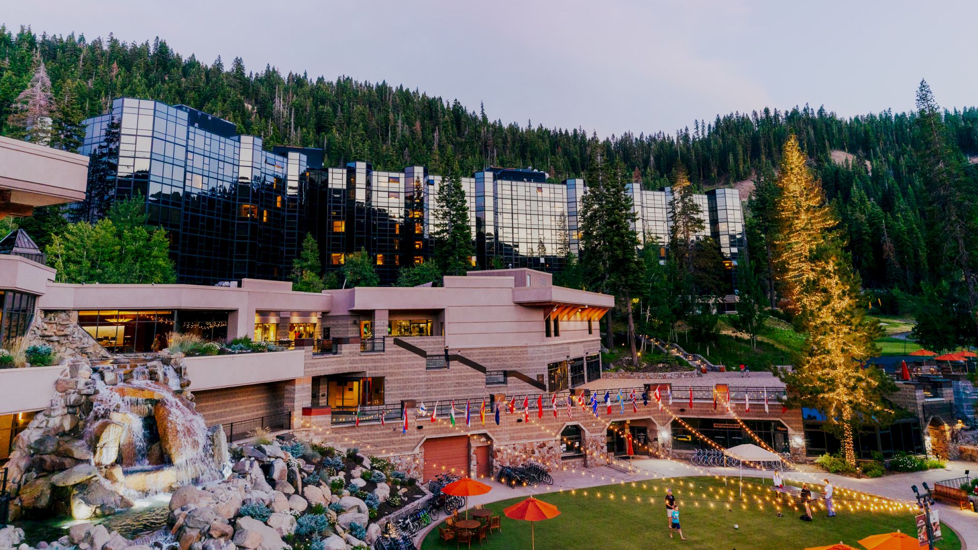 The image shows a modern resort with buildings featuring glass windows, surrounded by trees, a waterfall, and outdoor seating areas with string lights.