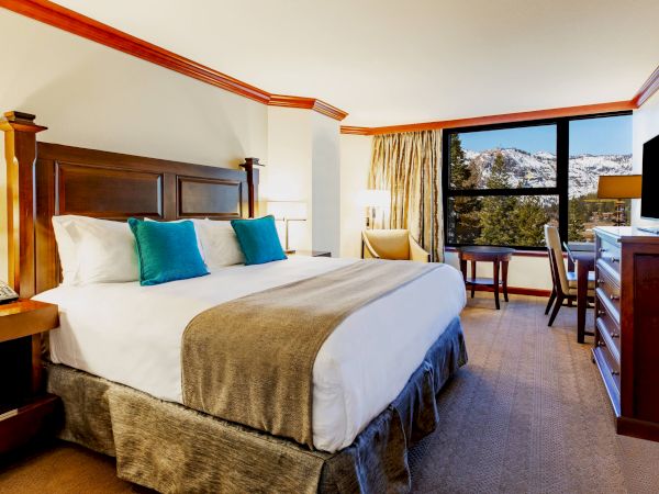 This image shows a hotel room with a large bed, wooden furniture, a television, and a window with a scenic view of a snowy mountain landscape.
