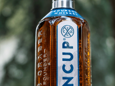 A bottle of Tito&rsquo;s/TINCUP straight bourbon whiskey with brown liquor, blue and white label, silver cap, outdoors blurred green background.